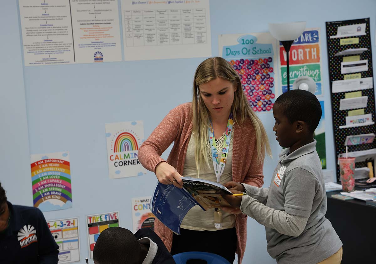 HSA Teacher smiles while kneeling beside a young student in a classroom setting.